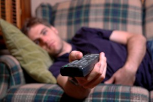 Weary looking young man lies on sofa extending a remote control.