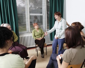 Woman standing, talking to seated women grouped in a circle, gesturing emotionally